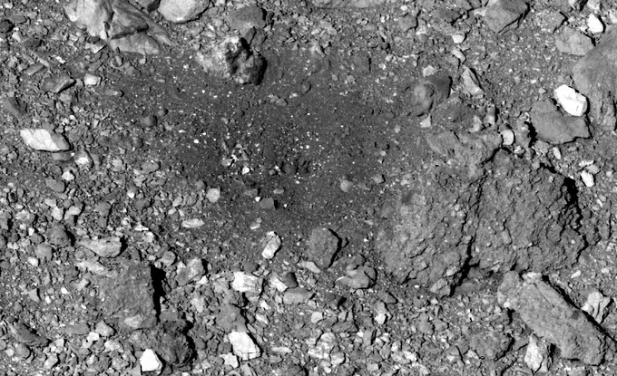 A black and white image of the spot where OSIRIS-REx captured the Bennu asteroid sample. The image features a close-up of a rocky rubble pile, with rocks of various sizes and shapes. Most of the rocks are roughly the same gray color as the background, but a few are a brighter white. In the center of the image, there is a relatively clear patch where the rocks are darker and smaller.