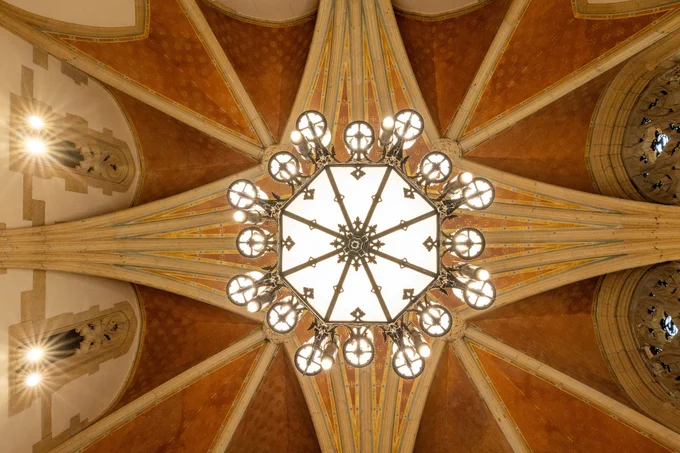 View from below of an elaborate chandelier hanging from the vaulted ceiling of a historical building, featuring intricate architectural details and warm lighting.