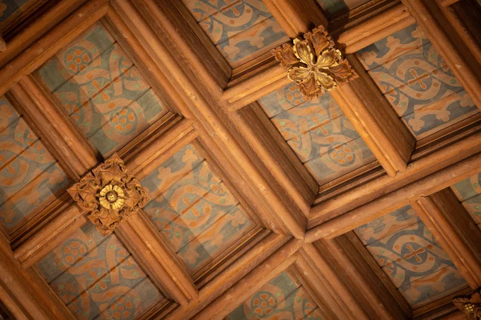 Decorative ceiling with intricate wooden beams featuring ornamental gold accents and patterns with crosses, possibly inside a historic building.
