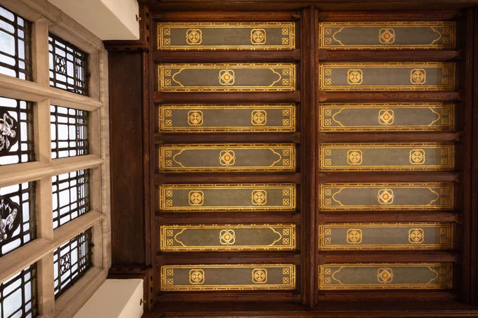 Interior view of an ornate wooden ceiling with geometric and floral designs, viewed from below, in a room with Tudor-style windows.