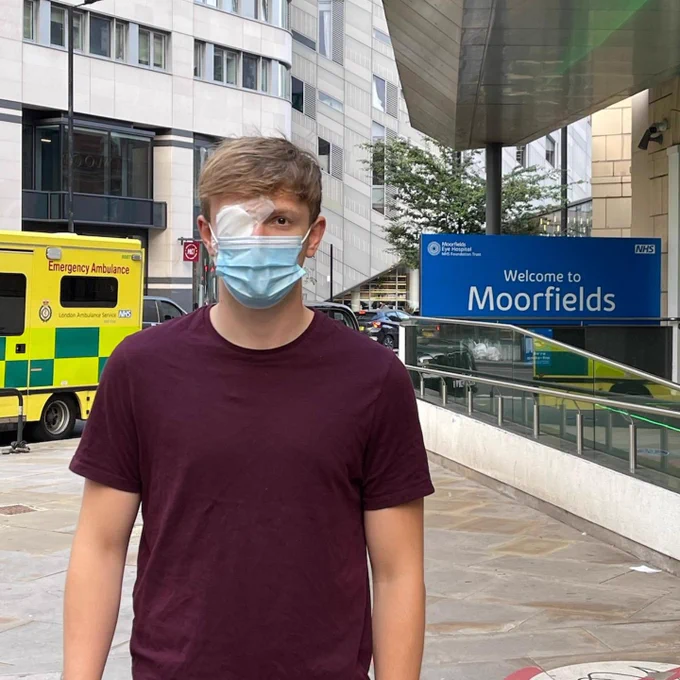 Myles standing outside the entrance to Moorfields Eye Hospital. He is wearing a medical eye patch and a face mask.