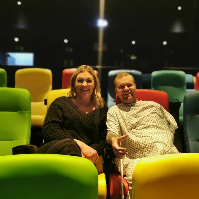 A man and woman smile for a picture inside a cinema. The cinema seats are green, orange, blue and yellow. The woman has blonde hair and is dressed in a black cable knit jumper, and is seated cross legged with her arms in her lap. The man is a patient and wears a hospital gown, with one arm tucked inside the gown, and the other resting on the seat armrest with his hand reaching slightly forward.