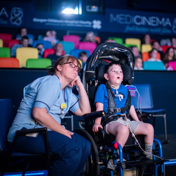 A female nurse and young male patient are watching a movie in a MediCinema cinema. The boy is in a wheelchair and is connected to specialist medical equipment. He dressed in a blue T-shirt and khaki shorts and is watching the film with wide eyes and is clinging to his seat. The nurse is wearing blue scrubs, black framed glasses and is leaning in to the boy, resting her chin on her fist on the arm of the wheelchair. The background is blurry but you can see colourful cinema seats with other guests watching the film