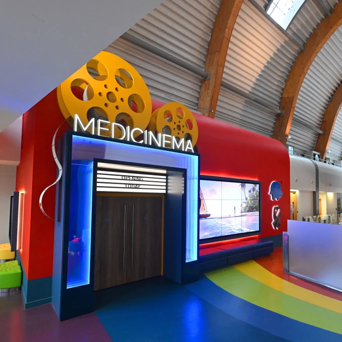 A MediCinema colourful entryway. The door entry is neon full and the double doors are wooden. Above the blue reads MEDICINEMA is white capital neon. Above the entry way a decorative oversized yellow film role and the floor is rainbow stripes of navy blue, blue, lime green, yellow and red. The cinema is located inside a large hospital, with a high ceiling made of corrugated iron and wooden beams.
