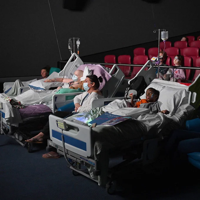 Two young patients in hospital beds are watching a film inside a cinema. They are both connected to IV drips are other specialised medical equipment. They are propped up in their beds and covered in white blankets. In between them a woman is seated and wearing a blue medical face mask. Her posture is relaxed and she too is watching the film. Behind the two young patients are two other patients. They are sitting in red cinema chairs and too are watching the film.