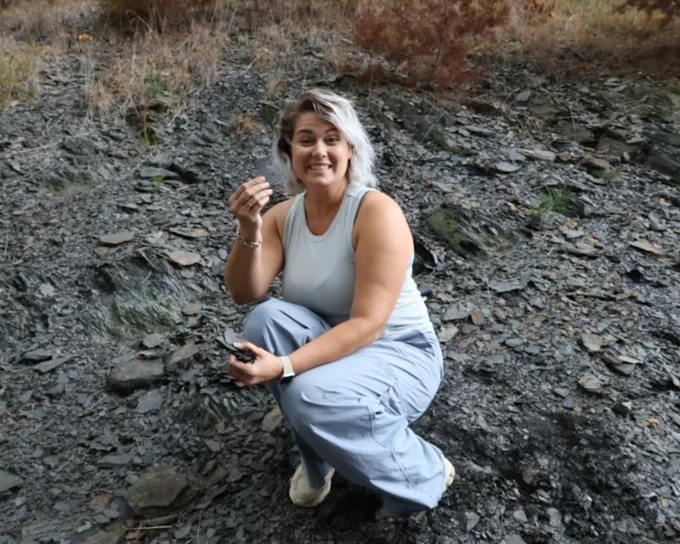 The student is half sitting in a field and smiling at the camera while holding a material. 