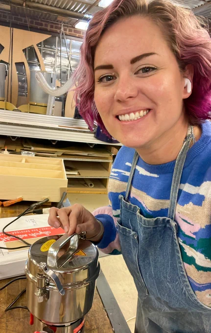 The student is working with certain tools in a lab. She is smiling at the camera. 