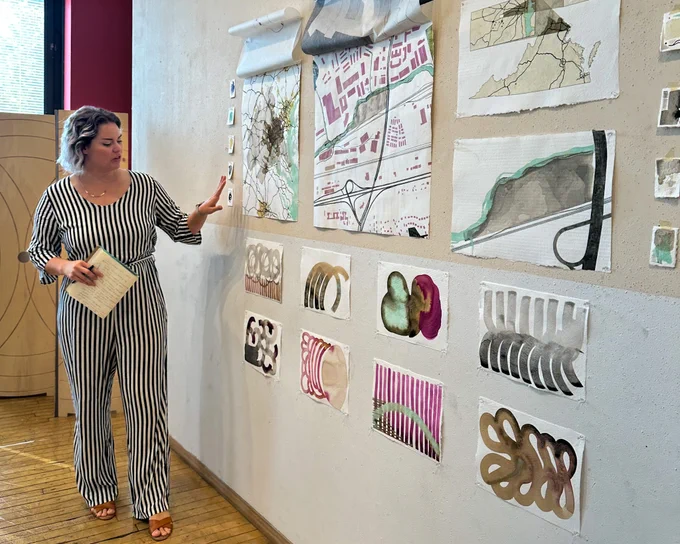 The student is standing in front of a wall with her work pinned up while she is pointing to her work and presenting. 