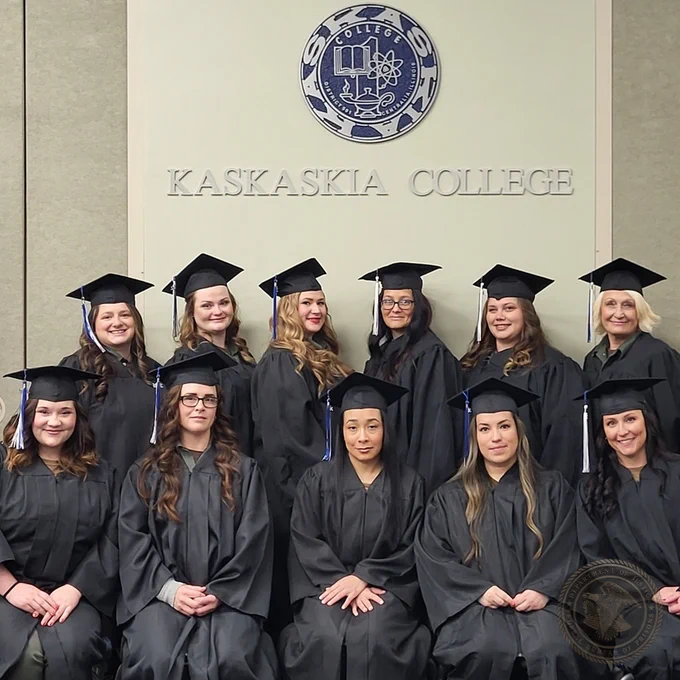 Group of graduates in caps and gowns posing in front of a Kaskaskia College seal and sign.