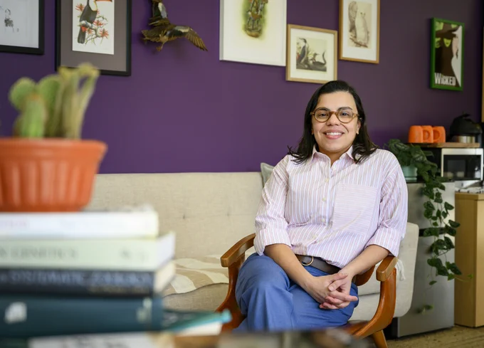 a smiling person with glasses is casually sitting in a chair in an office with stylish art and a deeply purple wall