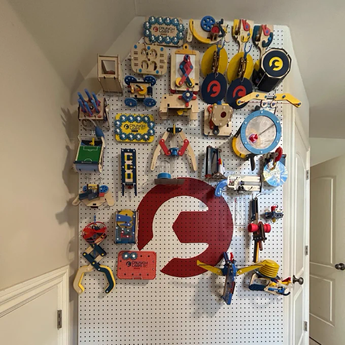 A wall-mounted organizer displaying an array of colorful wooden toys and gadgets, featuring a large red and white CrunchLabs logo in the center.
