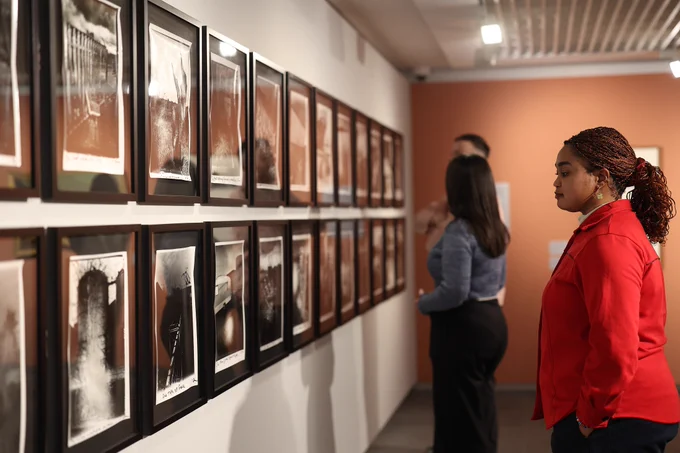 A photograph of three visitors examining a series of framed photographs in an art gallery. The person in the foreground is wearing a bright red jacket.