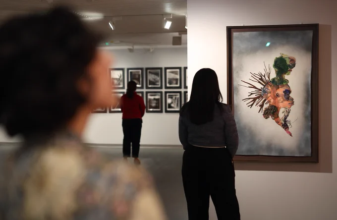 A photograph of three individuals viewing artworks in a gallery. The figure in the background is looking at framed photographs, while the figure in the middle is looking at a detailed and colourful abstract painting. The third figure is very close to the camera and blurred, whilst looking up.