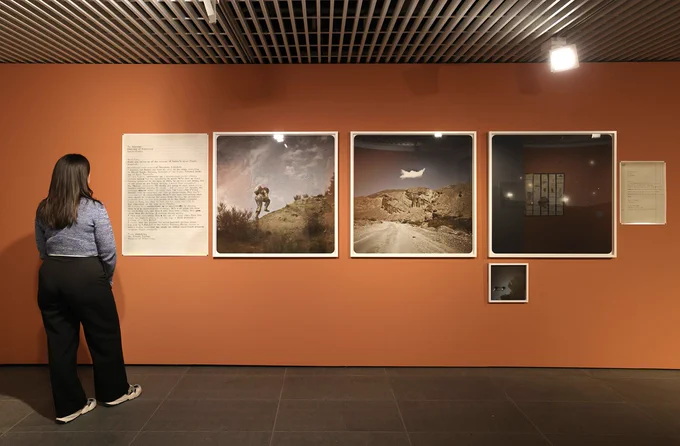 A photograph of a person viewing a series of photographs displayed on an orange gallery wall. 