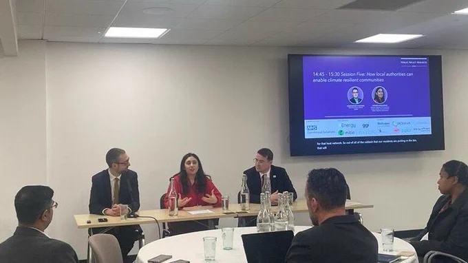 A panel discussion at the PPP Sustainable Estates Conference with speakers seated at a table, including Christopher Hammond from UK100, with an audience seated at round tables. A presentation screen shows the session title.