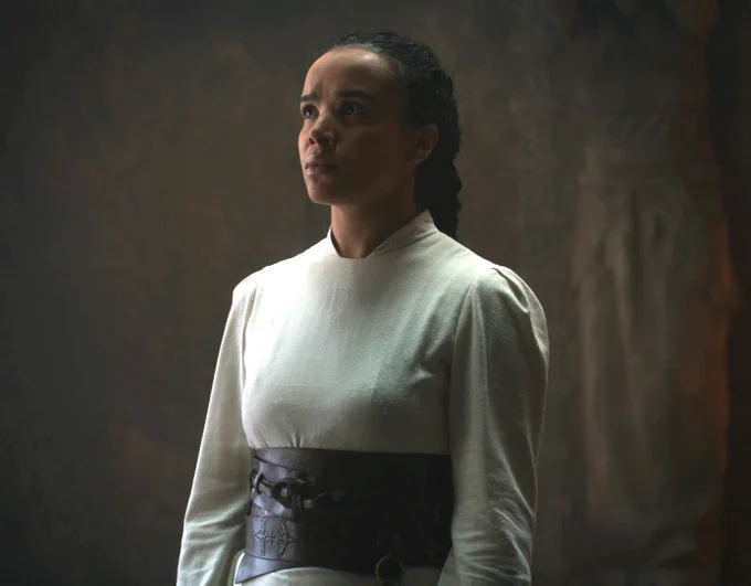 Zoë Robins as Nynaeve al'Meara. She has long dark hair pulled back in a braid. She wears a long-sleeved white dress with a wide brown leather belt.