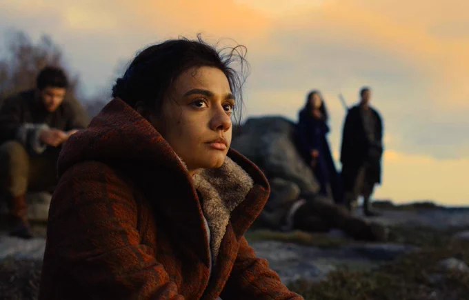 Madeleine Madden as Egwene al'Vere, sitting on a cliff. She has long dark brown hair. She wears a red-brown coat with white fur lining.
