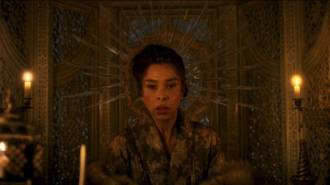 Sophie Okonedo as Siuan Sanche, seated in her carriage. She has short dark curly hair. She is wearing a green-and-gold kimono with a fur shawl. A halo of transparent blades hovers around her head.