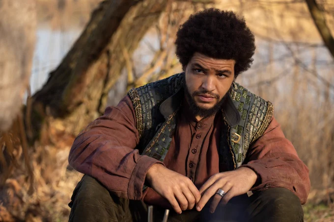Marcus Rutherford as Perrin Aybara, sitting by a riverbank. He has short dark frizzy hair and a beard. He is wearing a dark green woven-leather vest over a faded red long-sleeved shirt with olive-green trousers, and has a silver ring on his left hand.