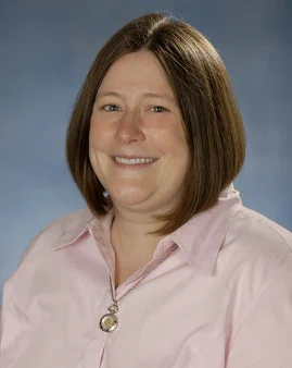 This image shows Dr. Julie Dunning Hotopp, Ph.D., sitting for a portrait in front of a blue-gray backdrop. She is wearing a pink collared shirt with a gold necklace and has brown hair cut into a bob. 