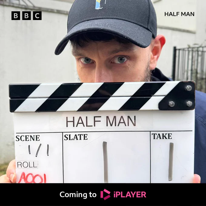 Richard Gadd holds a black and white clapper board in front of his face, only showing his eyes. He is wearing a blue shirt and a black cap. The board reads ‘ Half Man, Scene 1/1, Slate 1, Take 1.’ Text at the bottom reads ‘ Coming to iPlayer’. 