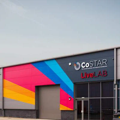 Exterior shot of a studio with bright pink, orange, blue and yellow lines painted on a grey exterior wall. The studio name is CoSTAR Live Lab