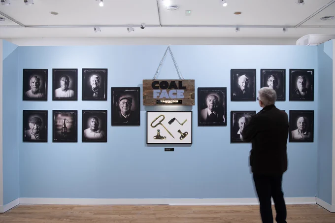 A pale blue wall in a bright gallery space. On the wall is a sign that reads 'Coal Face' and a selection of black and white tintype portrait photographs. A visitor with short grey hair, a dark top and dark blue jeans looks at the display.