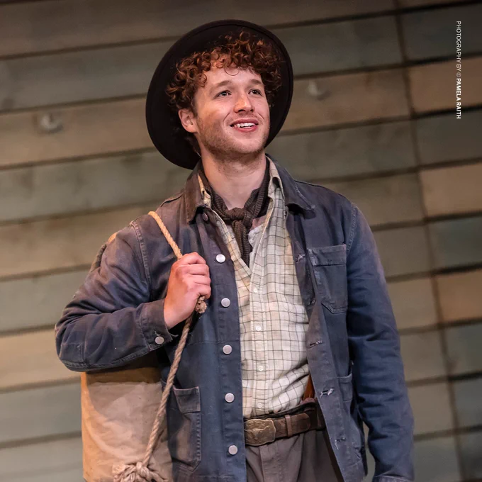 A young man (Liam King) with curly hair, dressed in a denim jacket, plaid shirt, and a wide-brimmed hat, smiles confidently while carrying a tied-up bedroll over his shoulder. The rustic wooden background suggests a rural setting. Photography credit: Pamela Raith