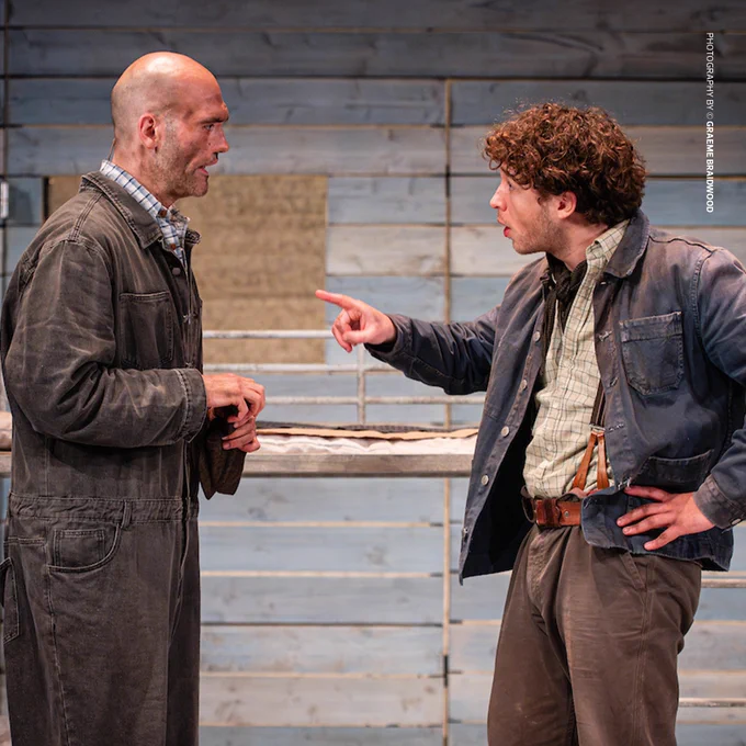 A tense moment between two male characters (Luke Richards - Left, Liam King - Right) in a bunkhouse setting, with wooden walls and metal bed frames in the background. The younger man, dressed in a denim jacket and suspenders, gestures forcefully at the bald, older man, who listens intently with a defensive stance. Photography Credit: Graeme Braidwood.