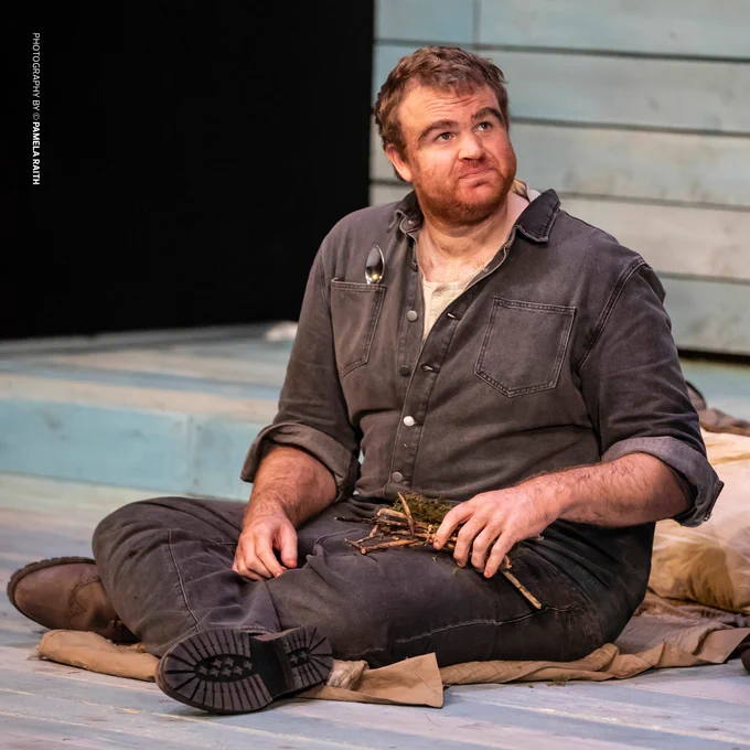 A bearded man with dishevelled hair, dressed in dark overalls, sits on the floor holding a small bundle of twigs. His expression is contemplative, and a spoon is clipped to his chest, emphasising a simple, working-class lifestyle. Photography Credit: Pamela Raith