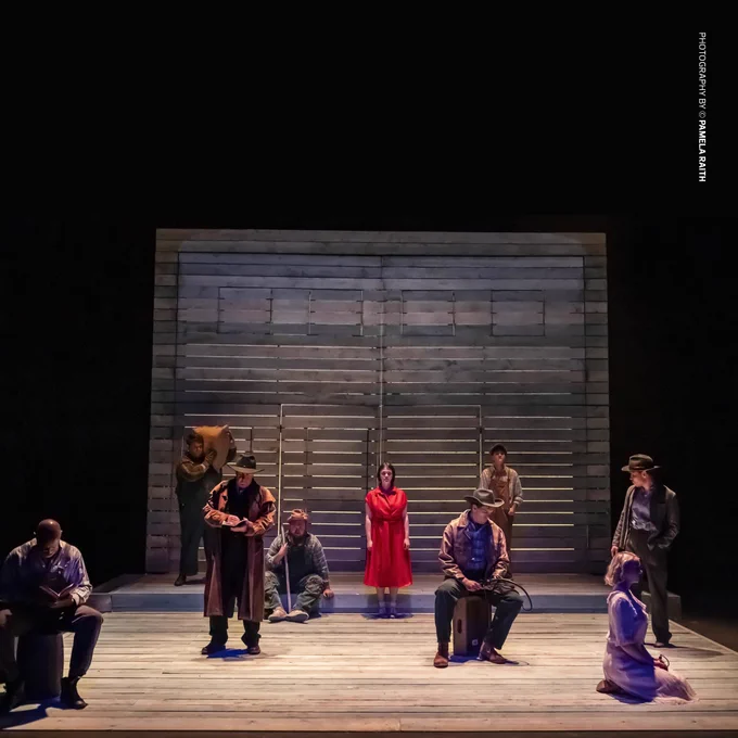 A stage performance of Of Mice and Men features a wooden set with slatted walls, dim lighting, and nine actors in period-appropriate 1930s attire. The central figure, a woman in a striking red dress, stands among male characters dressed as ranch workers and drifters, creating a dramatic tableau. Photography Credit: Pamela Raith