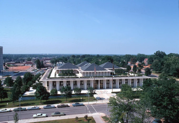 Aerial view of the North Carolina Legislative Building, featuring a large, white, modern structure with a flat roof, surrounded by spacious lawns and roads lined with parked cars.