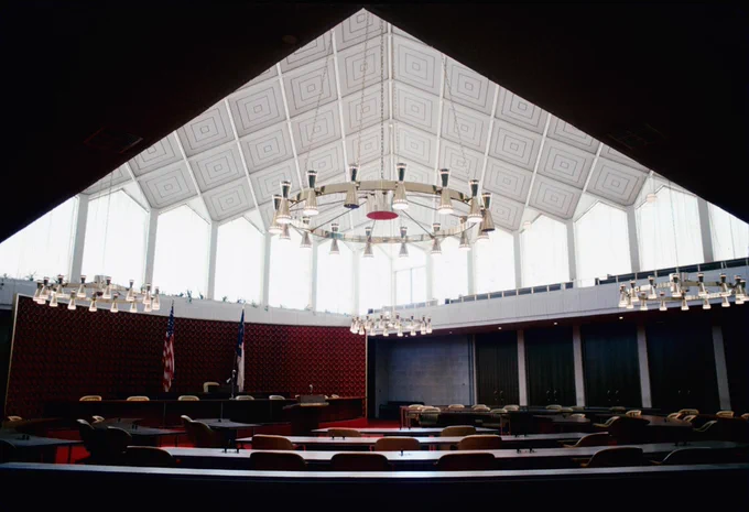 Interior view of the North Carolina Legislature chamber, featuring rows of desks, geometric ceiling design, and flags including the United States flag.