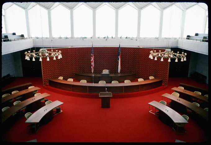Interior view of the North Carolina Legislature, featuring a semi-circular seating arrangement, red wall panels, and multiple rows of lights overhead.