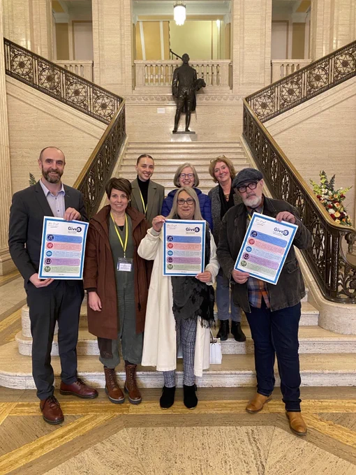 MLA Mark H Durkan and 6 New Script activists pose on the stairs in Stormont, 3 at the front holding ‘Give 5’ posters.