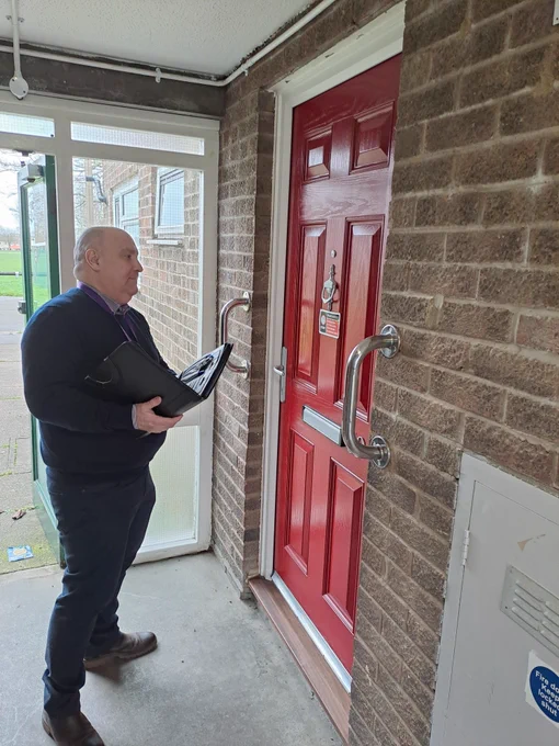 A man holding a folder stands outside a red door with accessibility grab bars, seemingly waiting for a response after knocking. The setting appears to be an apartment or housing complex with a covered walkway.