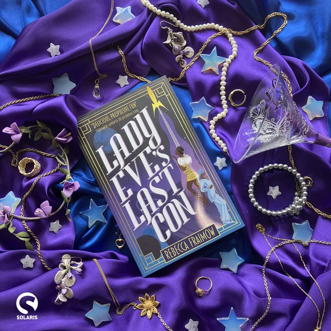 Against a background of purple and blue material dotted with jewellery, crystal stars and a purple cocktail glass, a paperback copy of LADY EVE'S LAST CON by Rebecca Fraimow. The white Solaris logo is in the bottom left corner.