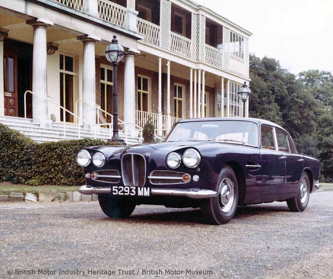 A colour photograph of a three-quarter view of a dark blue Aston Martin Lagonda Rapide parked outside of a building with a colonnade and lattice panelling. 