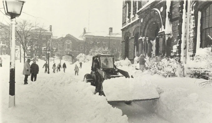 A historical black-and-white photo showing a snowplow clearing heavily snow-covered paths at Brown University, with people in the background. 