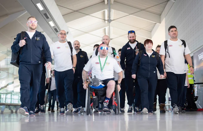 A group of people, mostly wearing Team UK white shirts and navy pants, walk through an airport. One person in a wheelchair is at the centre, wearing mirrored sunglasses. 