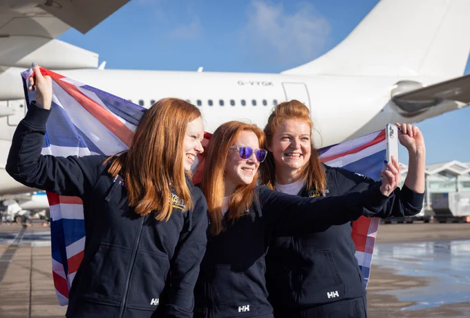 Three women in matching navy Team UK tracksuits take a selfie while holding a British flag. They stand in front of a large airplane on an airport tarmac. 