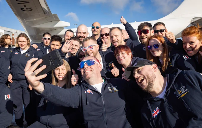 A group of people in matching Team UK uniforms pose enthusiastically for a selfie in front of an airplane. Many wear sunglasses, smiling and giving thumbs up to the camera.