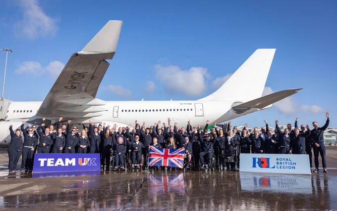 A group of people in uniform poses cheerfully in front of a large airplane on a sunny day. They hold a British flag. Signs read Team UK and Royal British Legion. The ground is wet, reflecting the blue sky and clouds.