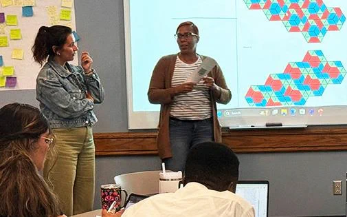Educators attend a professional learning presentation on computational thinking. Two adults sit at a table listening to two instructors standing in front of a white board showing math problems.