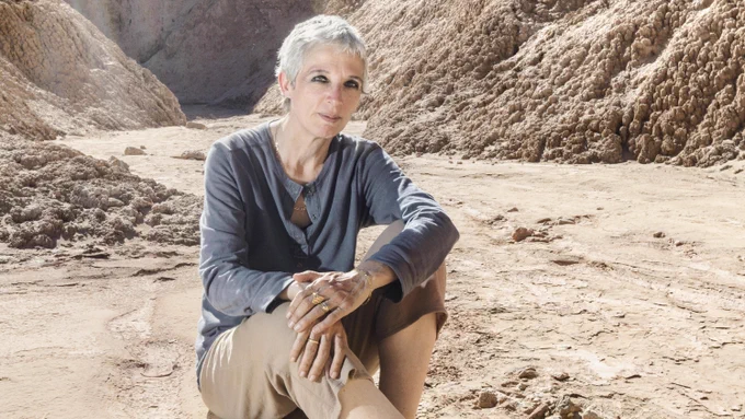 A woman with short white hair sits on a bed of sand and rock in the desert with rock formations behind her. She's wearing a slate blue longsleeve shirt and khaki pants. 