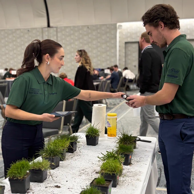Two individuals in branded shirts examine a small plant at an educational event.