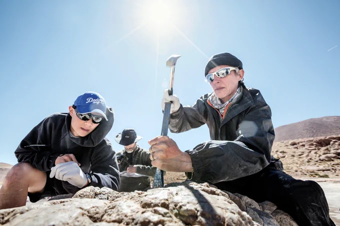 Three researchers in the desert take samples on a field expedition. A woman on the right is swinging a hammer to take a rock sample while her colleague on the left supervises, wearing a blue Dodgers baseball cap. The sun shines brightly overhead in a cloudless sky.