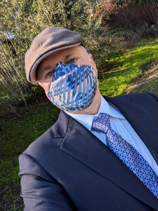 A man outdoors wearing a suit and tie, and a matching fabric mask cover over a 3M Aura 9210  n95.