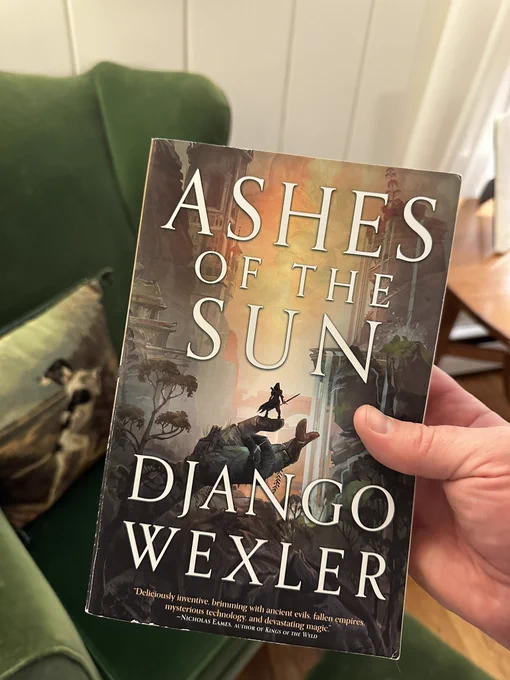 A hand holds a book called ASHES OF THE SUN, by Django Wexler, with a green velvet chair in the background. The cover depicts a lone figure holding a large sword standing in the thumb of a giant, ruined statue’s outstretched hand. A wilderness of ruins and a waterfall stretches ahead in a dramatic valley.