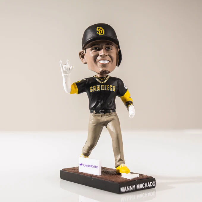 Photo of a Manny Machado bobblehead against a white backdrop. The bobblehead shows Manny holding up a "love" sign with one hand as he crosses home plate. He is wearing one of the Padres road uniforms (brown top and sand pants).